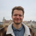 Headshot of Jonas Stettner, smiling, wearing a blue shirt and brown sherpa-collared jacket, with blurred historic buildings in the background.